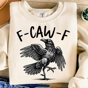 May include: Cream-colored sweatshirt with the text "F-CAW-F" above a detailed black and white illustration of a crow with outstretched wings. The crow is depicted in a dynamic pose.