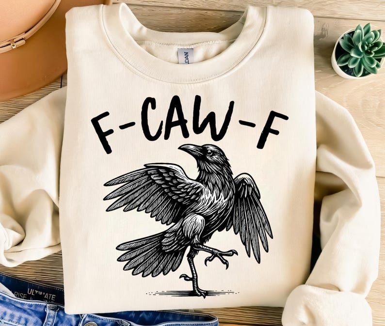 May include: Cream-colored sweatshirt with the text "F-CAW-F" above a detailed black and white illustration of a crow with outstretched wings. The crow is depicted in a dynamic pose.