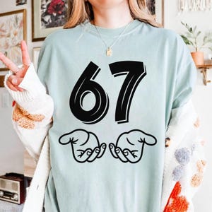 May include: A pale blue t-shirt featuring the number "67" in a thick black font. Beneath the numbers are two stylised hands with outstretched fingers. The shirt has a relaxed fit.