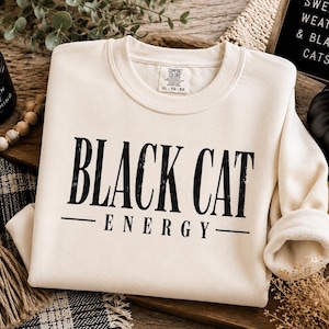 May include: A cream-colored crewneck sweatshirt featuring the black text "BLACK CAT ENERGY" printed on the front. The garment has a ribbed collar and long sleeves, with a visible interior tag indicating size XL. The design uses a bold, serif font.