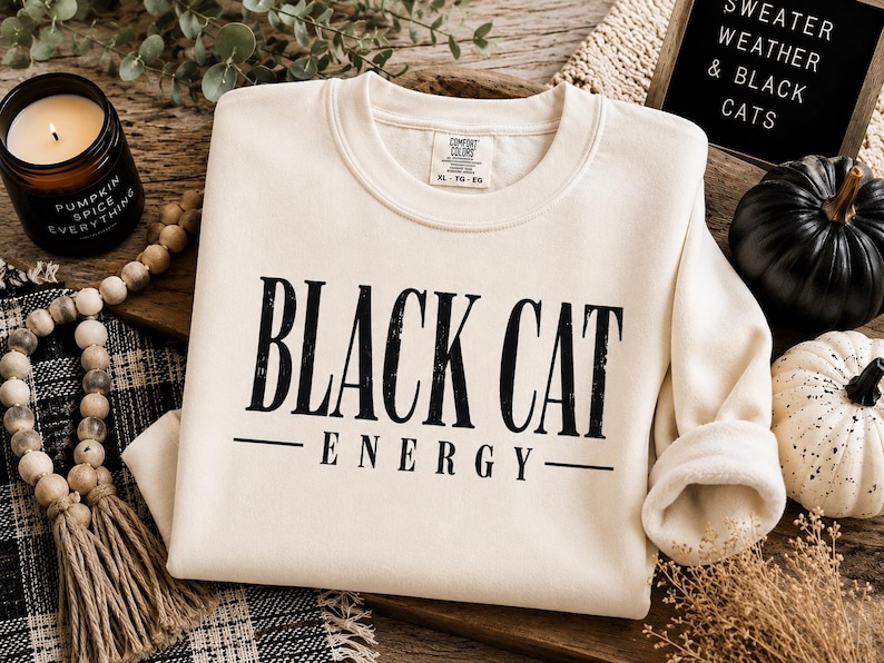 May include: A cream-colored crewneck sweatshirt featuring the black text "BLACK CAT ENERGY" printed on the front. The garment has a ribbed collar and long sleeves, with a visible interior tag indicating size XL. The design uses a bold, serif font.