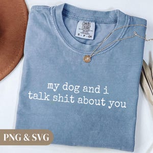 May include: A blue Comfort Colors t-shirt with the text "my dog and i talk shit about you" in white. A gold necklace with a pendant is draped across the shirt. The shirt has a tag that says "Comfort Colors".