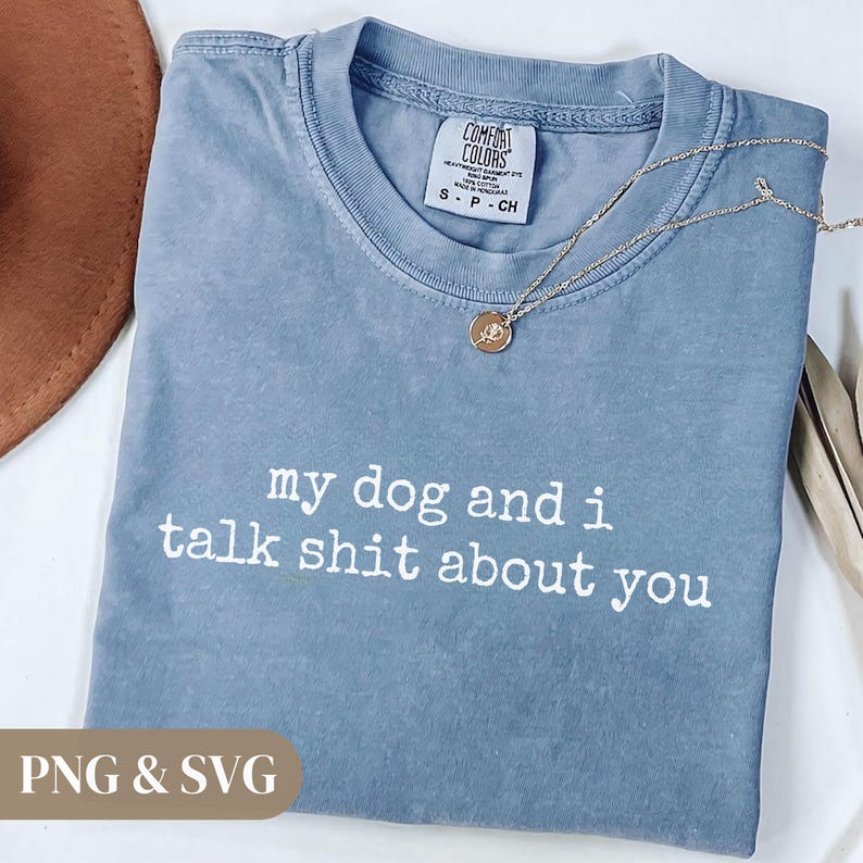 May include: A blue Comfort Colors t-shirt with the text "my dog and i talk shit about you" in white. A gold necklace with a pendant is draped across the shirt. The shirt has a tag that says "Comfort Colors".