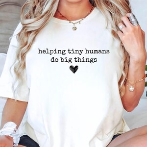May include: A cream-colored short-sleeve t-shirt featuring the black text "helping tiny humans do big things" printed on the front, centered above a small, hand-drawn style black heart icon. The shirt has a crew neckline and a relaxed fit.