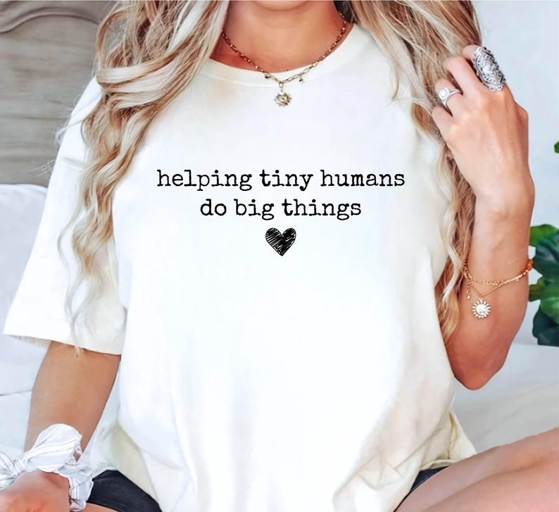 May include: A cream-colored short-sleeve t-shirt featuring the black text "helping tiny humans do big things" printed on the front, centered above a small, hand-drawn style black heart icon. The shirt has a crew neckline and a relaxed fit.