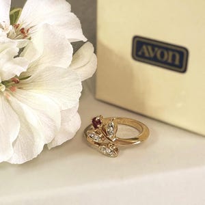 May include: A gold-tone ring featuring a small red stone and three marquise-shaped clear stones arranged in a leaf-like pattern. The ring is positioned next to a white flower and a cream-colored box with the text AVON printed in a dark blue rectangle.