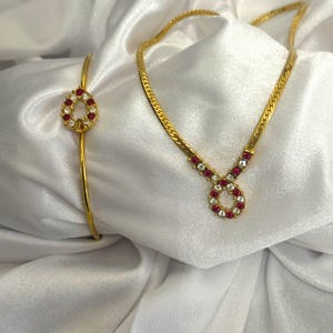 May include: A gold-toned jewelry set featuring a bracelet and necklace. The bracelet is a simple band with a circular pendant. The necklace has a chain with a matching circular pendant. Both pendants are adorned with red and clear gemstones.