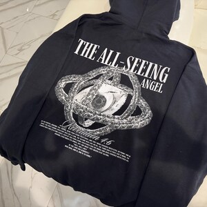 May include: Black hooded sweatshirt with a graphic on the back. The graphic features an eye within a circular design, with the text "THE ALL-SEEING ANGEL" above and "Revelation 4:6" below. Additional text is also present.