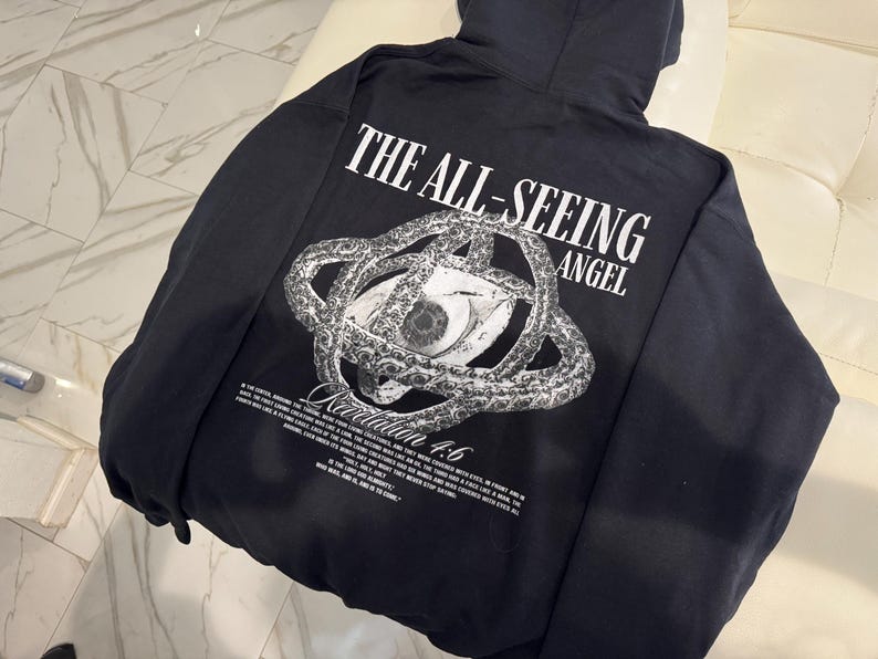 May include: Black hooded sweatshirt with a graphic on the back. The graphic features an eye within a circular design, with the text "THE ALL-SEEING ANGEL" above and "Revelation 4:6" below. Additional text is also present.
