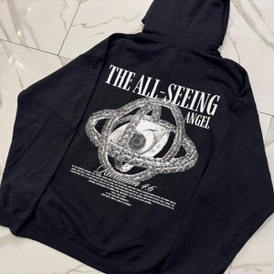May include: Black hooded sweatshirt with a graphic on the back. The graphic features an eye within a circular design, with the words "THE ALL-SEEING ANGEL" above it. Below the graphic is the text "Revelation 4:6".