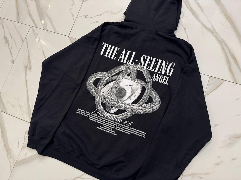 May include: Black hooded sweatshirt with a graphic on the back. The graphic features an eye within a circular design, with the words "THE ALL-SEEING ANGEL" above it. Below the graphic is the text "Revelation 4:6".