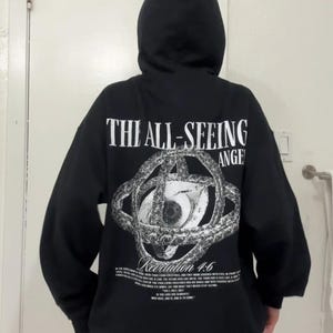 May include: Black hooded sweatshirt with the text "THE ALL-SEEING ANGEL" and an eye graphic on the back. The graphic includes the text "Revelation 4:6" and additional text below. The sweatshirt has a drawstring hood.