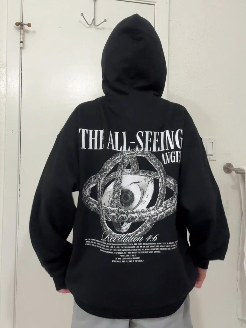 May include: Black hooded sweatshirt with the text "THE ALL-SEEING ANGEL" and an eye graphic on the back. The graphic includes the text "Revelation 4:6" and additional text below. The sweatshirt has a drawstring hood.