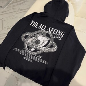 May include: Black hooded sweatshirt with the text "THE ALL-SEEING ANGEL" and an eye graphic. The graphic is surrounded by a serpent-like design. The text "Revelation 4:6" is also visible.
