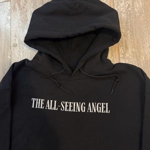 May include: Black hooded sweatshirt with the text "THE ALL-SEEING ANGEL" in white block letters. The sweatshirt has a drawstring hood and a kangaroo pocket. The fabric appears to be a soft, medium-weight material.