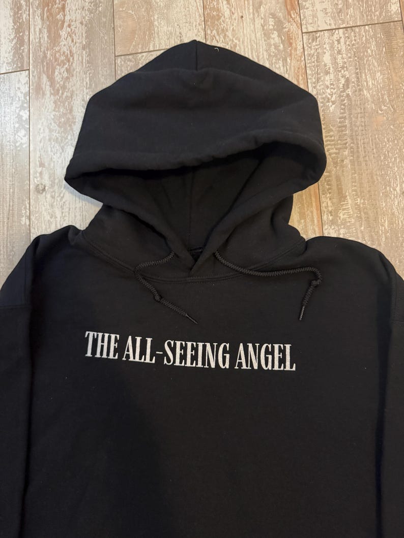 May include: Black hooded sweatshirt with the text "THE ALL-SEEING ANGEL" in white block letters. The sweatshirt has a drawstring hood and a kangaroo pocket. The fabric appears to be a soft, medium-weight material.