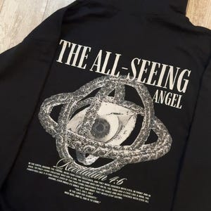 May include: Black hoodie with the text "THE ALL-SEEING ANGEL" and a graphic of an eye within a circular design. The text "Revelation 4:6" is below the graphic, along with a Bible verse.