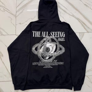 May include: Black hooded sweatshirt with the text "THE ALL-SEEING ANGEL" above a detailed eye graphic. Below the graphic is the text "Revelation 4:6" and other text. The sweatshirt has long sleeves and a hood.