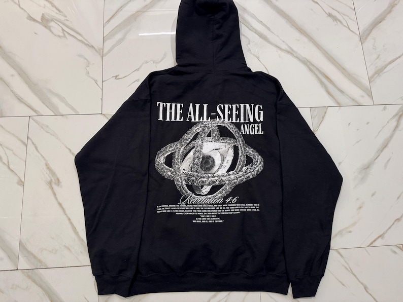 May include: Black hooded sweatshirt with the text "THE ALL-SEEING ANGEL" above a detailed eye graphic. Below the graphic is the text "Revelation 4:6" and other text. The sweatshirt has long sleeves and a hood.