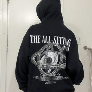 May include: Black hooded sweatshirt with the text "THE ALL-SEEING" above a detailed eye graphic. The graphic is surrounded by a chain-like design. Below the graphic is the text "Revelation 4:6" and additional text.