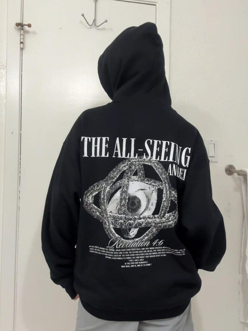 May include: Black hooded sweatshirt with the text "THE ALL-SEEING" above a detailed eye graphic. The graphic is surrounded by a chain-like design. Below the graphic is the text "Revelation 4:6" and additional text.