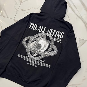 May include: Black hoodie featuring the text "THE ALL-SEEING ANGEL" above a detailed eye graphic. The design includes a complex, circular pattern around the eye and additional text below. The hoodie has a hood.