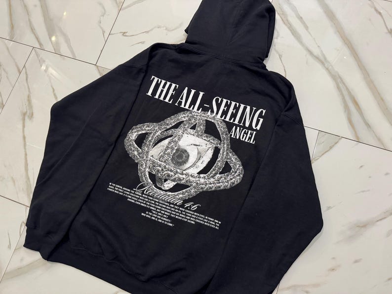 May include: Black hoodie featuring the text "THE ALL-SEEING ANGEL" above a detailed eye graphic. The design includes a complex, circular pattern around the eye and additional text below. The hoodie has a hood.