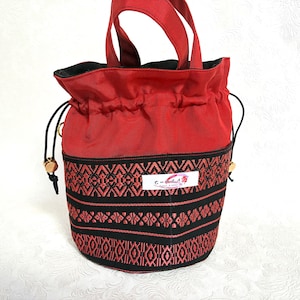 May include: Red drawstring bucket bag with black and red patterned fabric detail. The bag has a black interior and red handles. The bag has a small white tag with text and a logo.