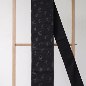 May include: A long, rectangular black sash featuring a repeating pattern of subtle, dark gray calligraphic symbols. The fabric has a smooth, slightly reflective finish, and the design is consistent across the entire length of the textile accessory.