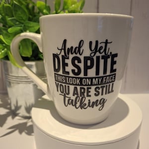 Puede incluir: Taza de cerámica blanca con texto negro: "And Yet, DESPITE THIS LOOK ON MY FACE YOU ARE STILL talking." La taza tiene un asa curva y está sobre una plataforma blanca. Una pequeña planta en maceta está en el fondo.