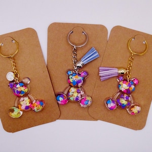 May include: Three clear acrylic bear keyrings filled with colourful confetti. Each keyring has a metal ring and chain. One has a gold ring, the other two have silver rings. Two keyrings have a tassel.