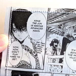 May include: A rectangular wallet featuring a black and white manga print with speech bubbles containing the text: Kang Tae-Jun is making advances again. Now, now. Don't play hard to get. Her face is so red. She's really trying hard. I'm not going to the imperial villa with you. Let go of me.