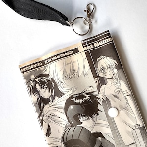 May include: A black and white manga-style wallet with a black strap and silver clasp. The wallet features manga illustrations and text that reads "MISSION-30 At the end of the battle" and "Bet Memo".