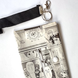 May include: A black and white manga-themed wallet with a black wrist strap and silver-colored clasp. The wallet features panels of manga art with Japanese text. The wrist strap is made of a woven material.