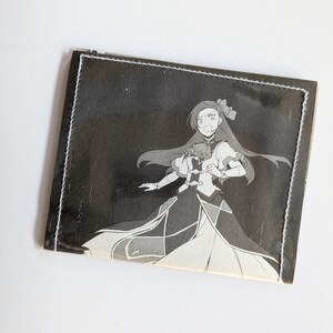 Puede incluir: Una cartera de cuero negro con un borde cosido blanco. La cartera presenta una ilustraci&oacute;n en blanco y negro de un personaje de anime con cabello largo y un vestido.
