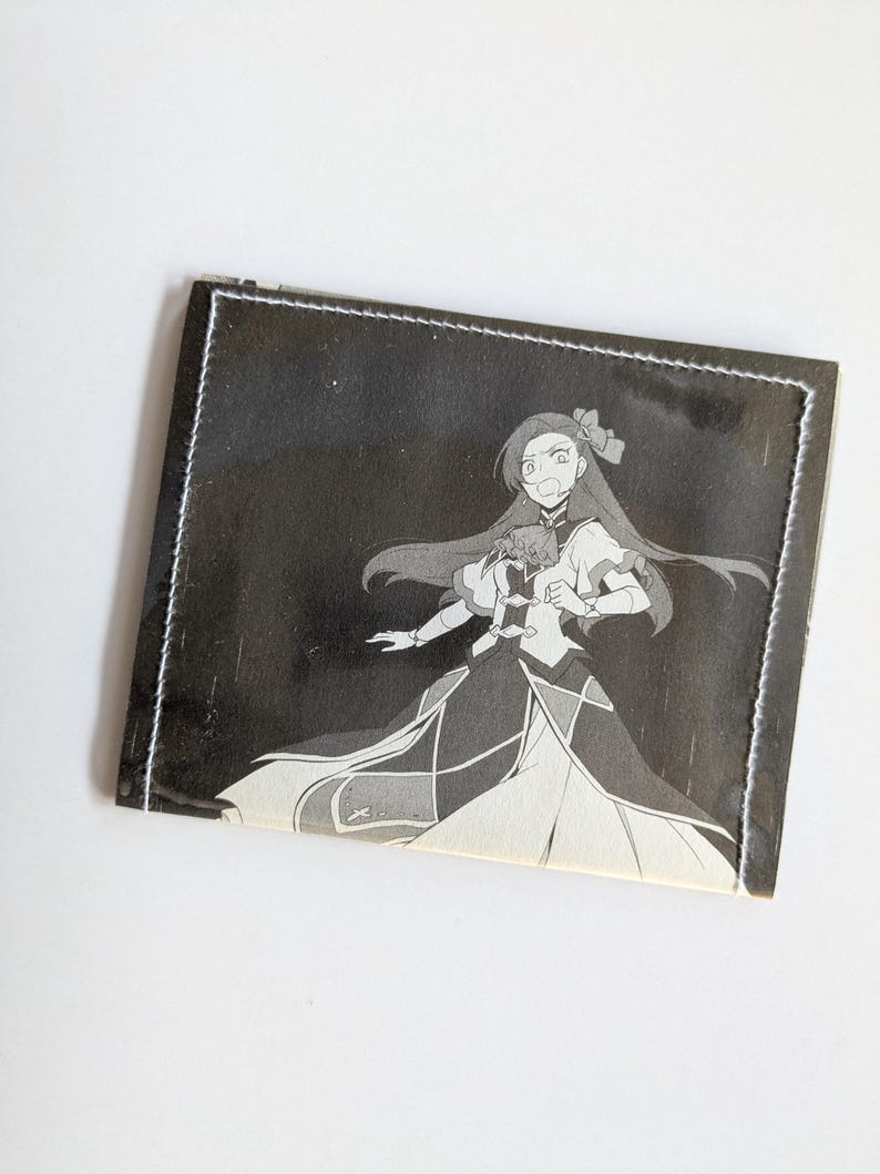 Puede incluir: Una cartera de cuero negro con un borde cosido blanco. La cartera presenta una ilustraci&oacute;n en blanco y negro de un personaje de anime con cabello largo y un vestido.