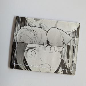 Puede incluir: Una cartera en blanco y negro hecha de un c&oacute;mic manga. La cartera presenta un primer plano de la cara de un personaje con ojos grandes y la boca abierta.