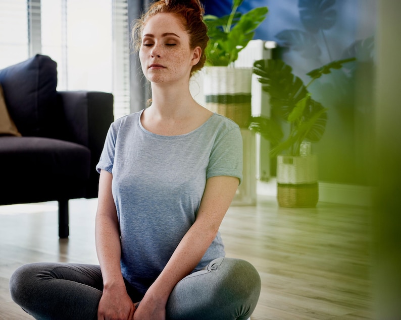 Puede incluir: Una persona con una camiseta azul claro y leggings grises est&aacute; sentada con las piernas cruzadas en un suelo de madera, con los ojos cerrados en meditaci&oacute;n. La habitaci&oacute;n cuenta con un sof&aacute; gris oscuro, plantas y luz natural.