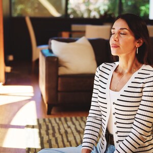 Puede incluir: Una persona con una camiseta sin mangas blanca y una rebeca a rayas blancas y negras. Est&aacute; sentada con los ojos cerrados, con un collar y pendientes. El fondo incluye un sof&aacute;, una alfombra y una ventana.