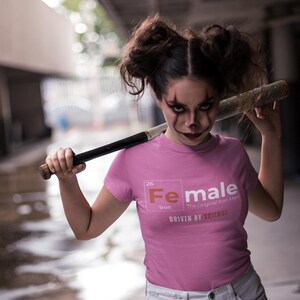 Puede incluir: Una camiseta rosa con un gr&aacute;fico blanco del elemento hierro de la tabla peri&oacute;dica, con la palabra "Female" en lugar del nombre del elemento. El texto "The Original Iron Man" y "Driven by Science" tambi&eacute;n est&aacute;n incluidos en el gr&aacute;fico.