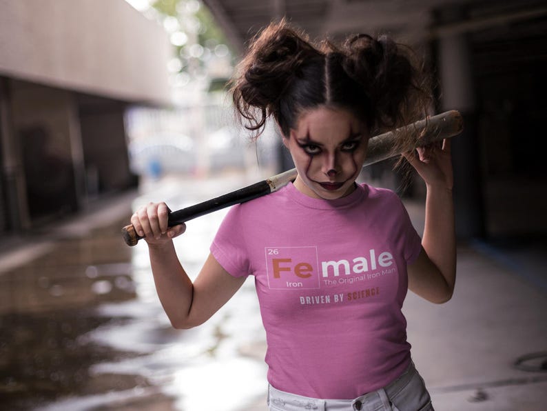 Puede incluir: Una camiseta rosa con un gr&aacute;fico blanco del elemento hierro de la tabla peri&oacute;dica, con la palabra "Female" en lugar del nombre del elemento. El texto "The Original Iron Man" y "Driven by Science" tambi&eacute;n est&aacute;n incluidos en el gr&aacute;fico.