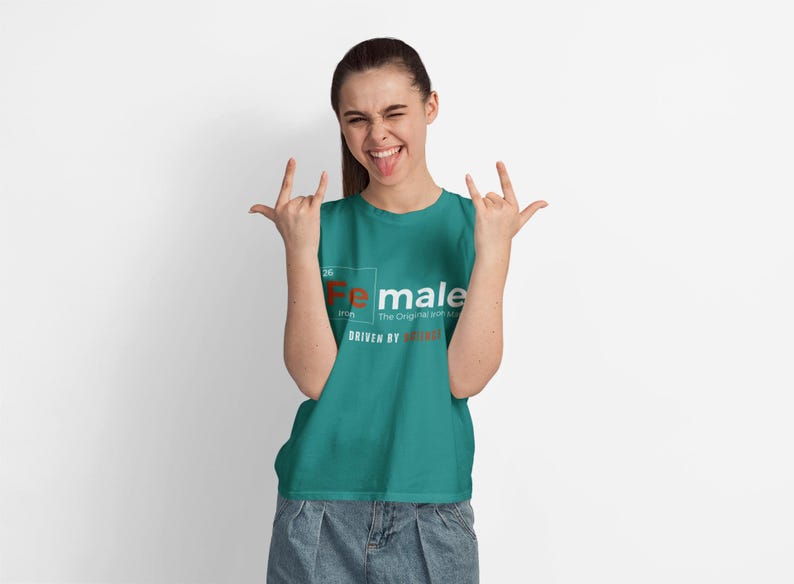 Puede incluir: Una camiseta azul turquesa con un gr&aacute;fico blanco que dice "Fe male" con la "Fe" en marr&oacute;n. El texto debajo dice "The Original and Still the Best" y "Driven by Strength".