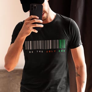 Puede incluir: Camiseta negra de cuello redondo con diseño de código de barras. El código de barras tiene rayas blancas, negras, rojas y verdes, con las palabras "BE THE ONLY ONE" impresas debajo. Una persona lleva la camiseta y una gorra negra.