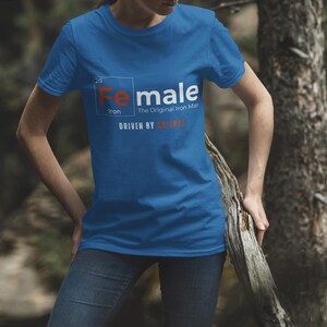 Puede incluir: Una camiseta azul con un gr&aacute;fico blanco de un elemento de la tabla peri&oacute;dica. El elemento est&aacute; etiquetado como "Fe" para el hierro y la palabra "Female" est&aacute; escrita sobre el s&iacute;mbolo del elemento. El texto "The Original Iron Man" est&aacute; escrito debajo del s&iacute;mbolo del elemento. El texto "Driven by" est&aacute; escrito debajo del s&iacute;mbolo del elemento y la palabra "Science" est&aacute; escrita debajo del texto "Driven by".