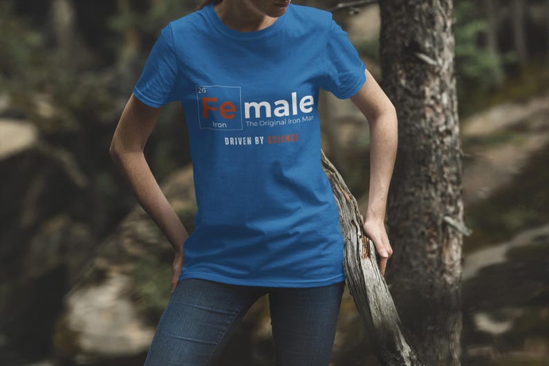 Puede incluir: Una camiseta azul con un gr&aacute;fico blanco de un elemento de la tabla peri&oacute;dica. El elemento est&aacute; etiquetado como "Fe" para el hierro y la palabra "Female" est&aacute; escrita sobre el s&iacute;mbolo del elemento. El texto "The Original Iron Man" est&aacute; escrito debajo del s&iacute;mbolo del elemento. El texto "Driven by" est&aacute; escrito debajo del s&iacute;mbolo del elemento y la palabra "Science" est&aacute; escrita debajo del texto "Driven by".