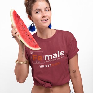 Puede incluir: Una mujer lleva una camiseta granate con un gr&aacute;fico blanco del elemento hierro de la tabla peri&oacute;dica, con la palabra "Female" escrita sobre el s&iacute;mbolo del elemento. El texto debajo del gr&aacute;fico dice "The Original Iron Man" y "Driven by Science".