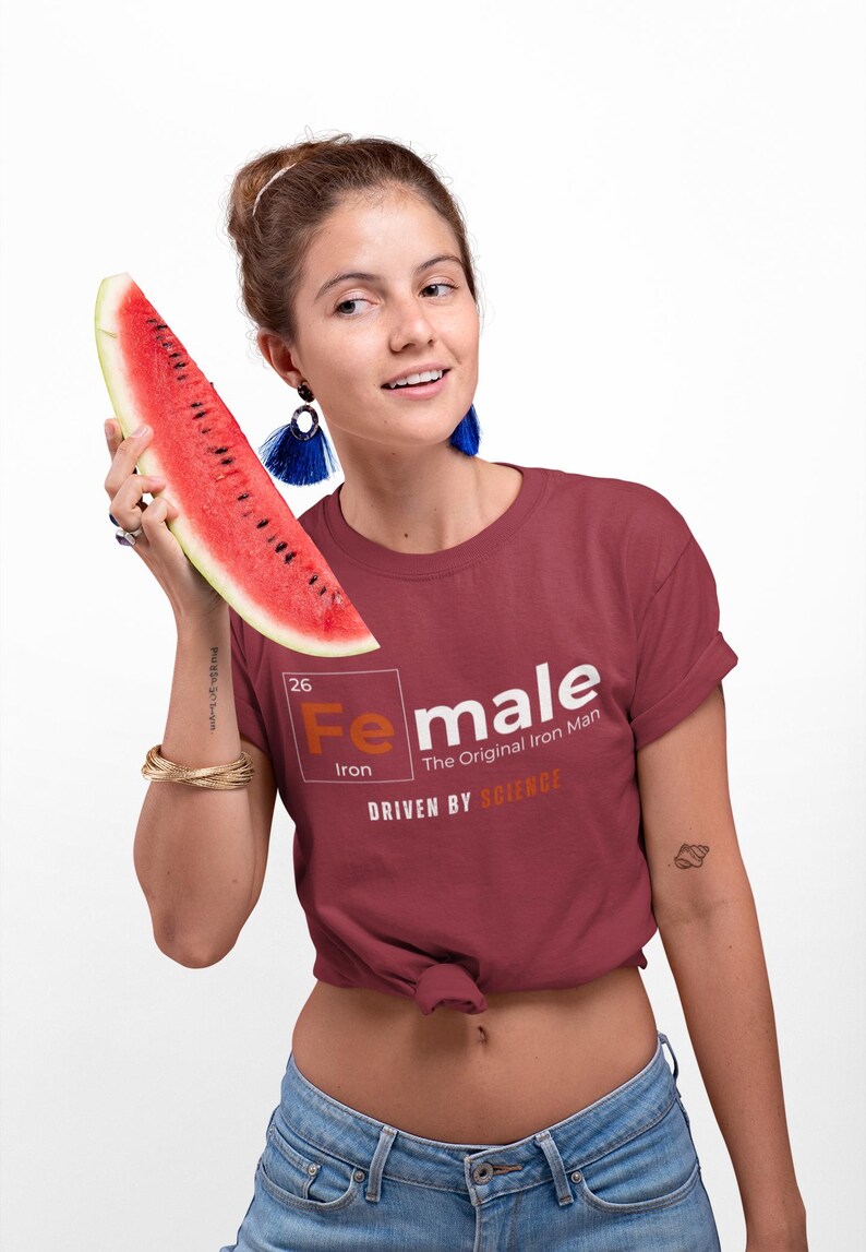 Puede incluir: Una mujer lleva una camiseta granate con un gr&aacute;fico blanco del elemento hierro de la tabla peri&oacute;dica, con la palabra "Female" escrita sobre el s&iacute;mbolo del elemento. El texto debajo del gr&aacute;fico dice "The Original Iron Man" y "Driven by Science".