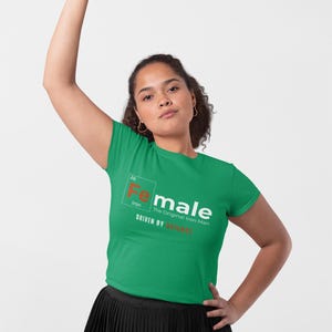 Puede incluir: Camiseta verde de manga corta con un gr&aacute;fico blanco de un elemento de la tabla peri&oacute;dica, "Fe" para el hierro, con la palabra "female" escrita a su lado. El texto debajo dice "The Original Iron Man" y "Driven by Instinct".