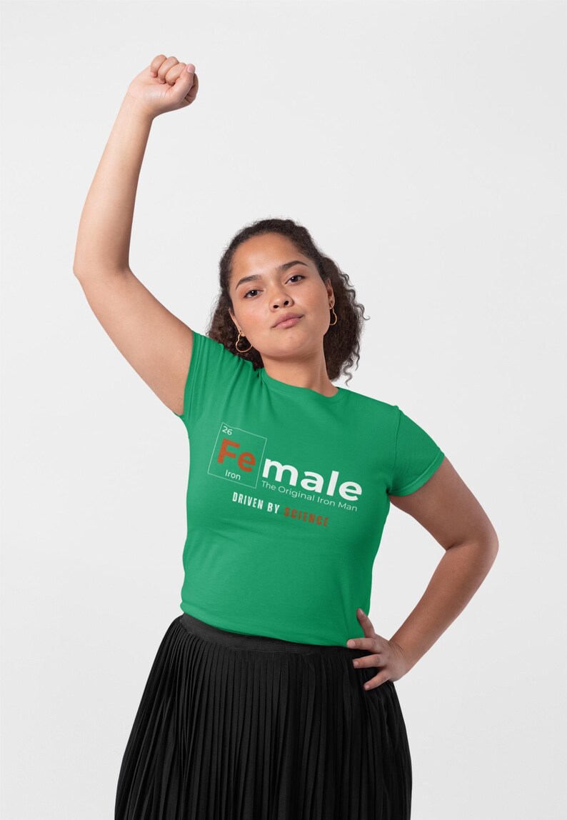 Puede incluir: Camiseta verde de manga corta con un gr&aacute;fico blanco de un elemento de la tabla peri&oacute;dica, "Fe" para el hierro, con la palabra "female" escrita a su lado. El texto debajo dice "The Original Iron Man" y "Driven by Instinct".