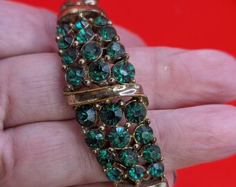 May include: Vintage gold-tone bracelet with three rows of emerald green rhinestones. The bracelet features a clasp closure and is set against a red background. The stones are round and faceted, adding sparkle to the piece. A classic jewelry item.